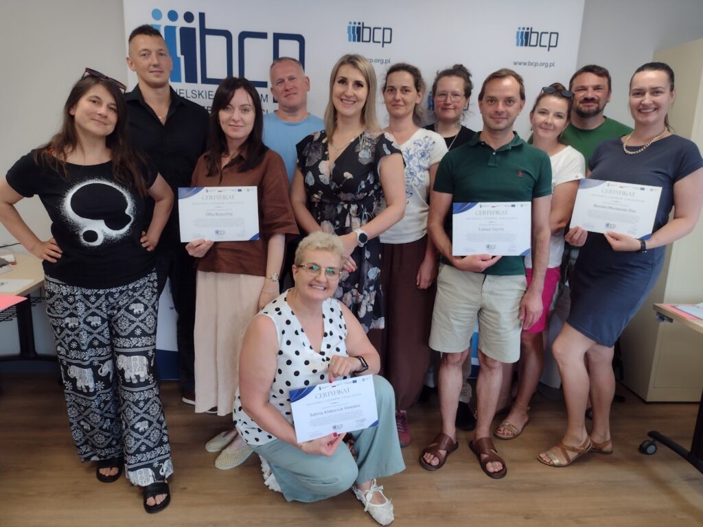 11 osób stoi na tle ścianki z logotypem BCP, jedna osoba kuca. Cztery osoby trzymają certyfikaty